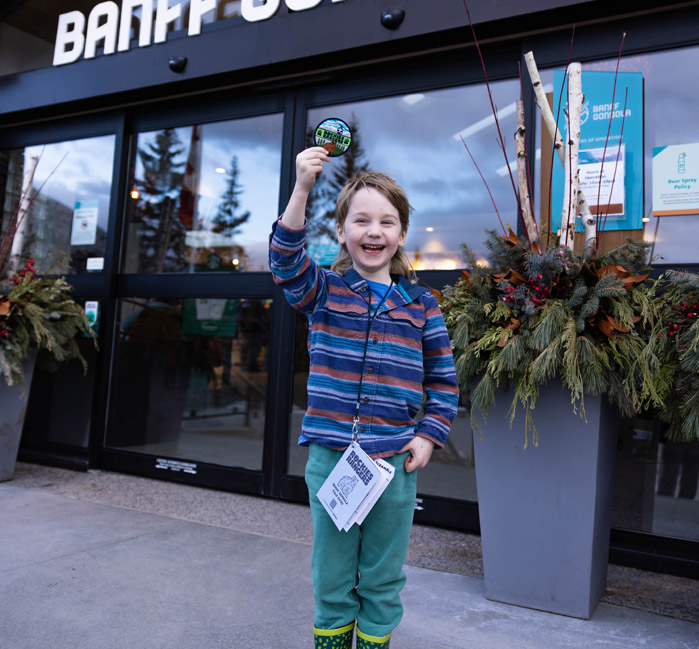 A kid smiling and proudly holding their Rockies Rangers badge up high while standing in front of the Banff Gondola entrance.