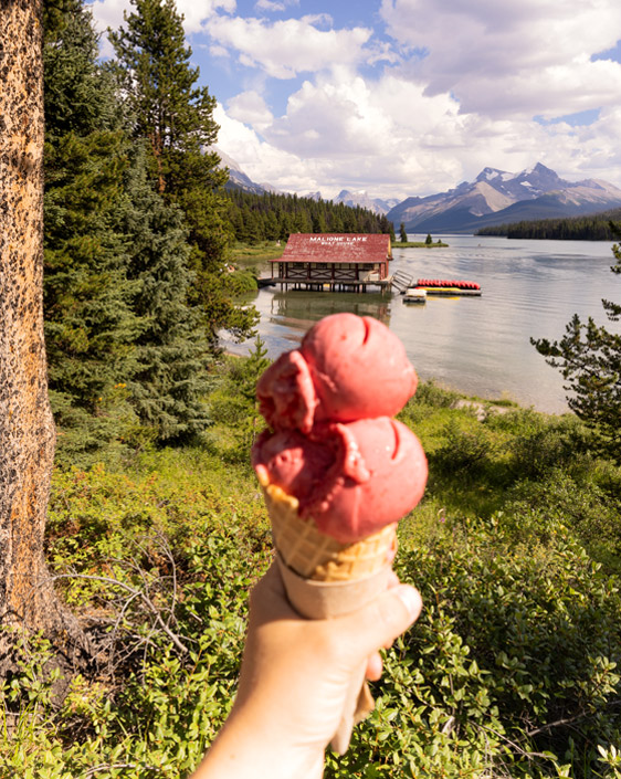 A person's hand holding an ice cream cone with Maligne Lake boat rentals and the mountains in the background.