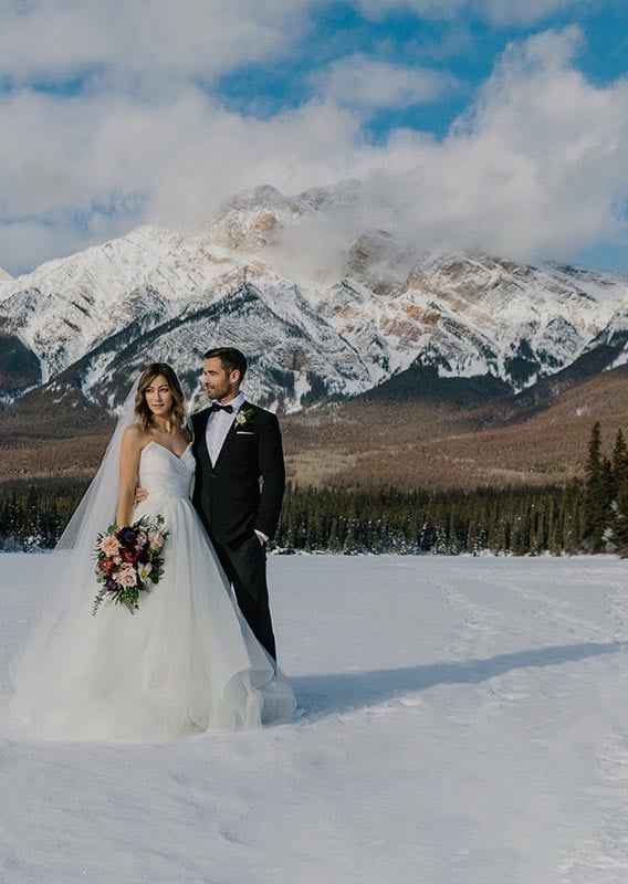 A newlywed couple stands on a snowy, frozen lake below blue skies and tall mountains.