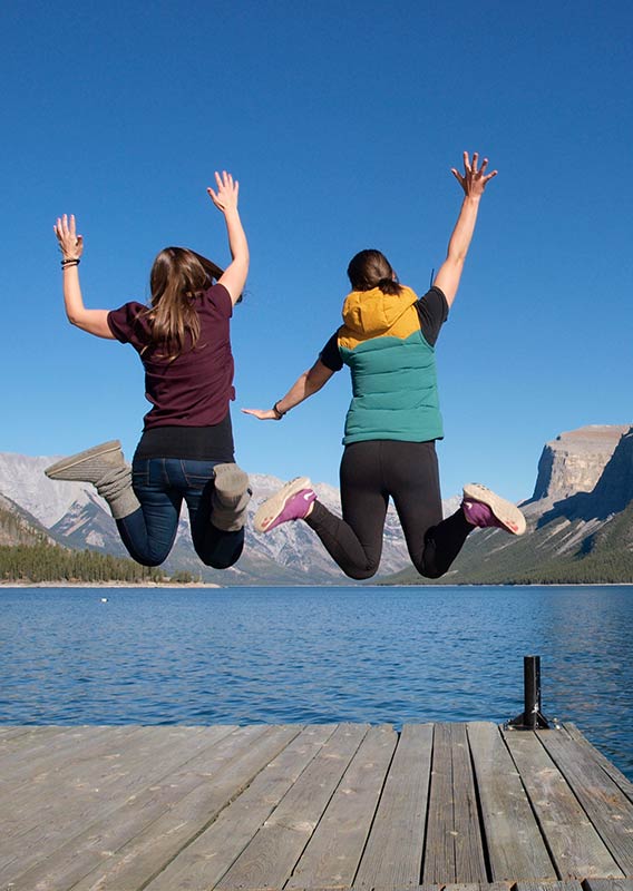 Two women jump in the air on a lake dock, with mountains in the background during summer.