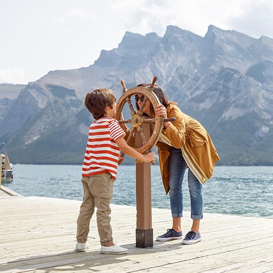 A parent and child look through a boat steering wheel mounted on a lakeside dock.
