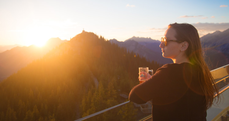 A person standing on the patio at the Banff Gondola summit looking at the sunset and mountains