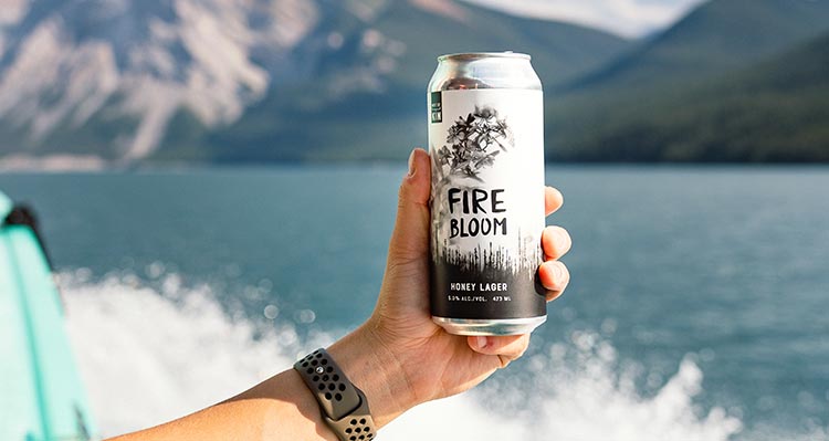 A can of Fire Bloom beer held up against a backdrop of mountains and a big lake.