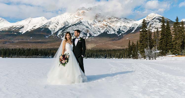 A bride and groom pose in the winter with a mountain behind them.