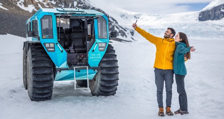 Two people taking a selfie together at Athabasca Glacier, with Ice Odyssey vehicle in the background.