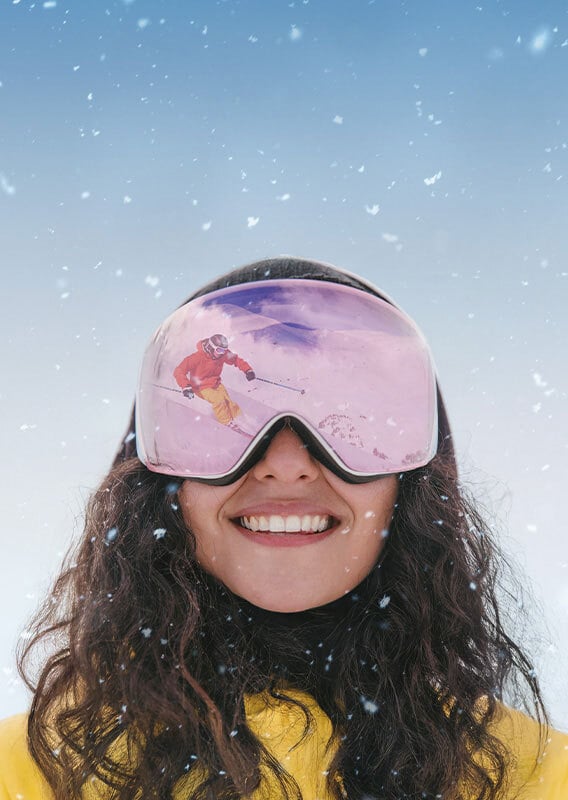 A woman smiles and a skiing scene is reflected in her goggles.