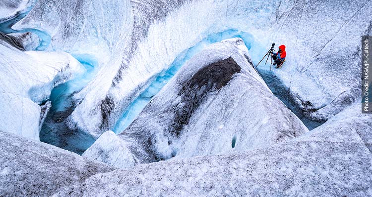 A researcher with a tripod crouches next to a winding stream of water in an icefield.