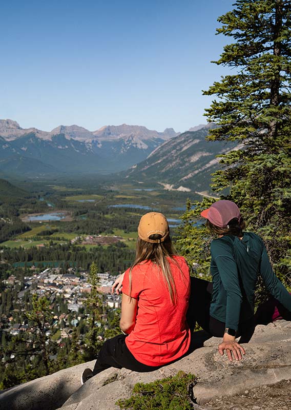 Two hikers sit on a rocky viewpoint above a small town below mountains.