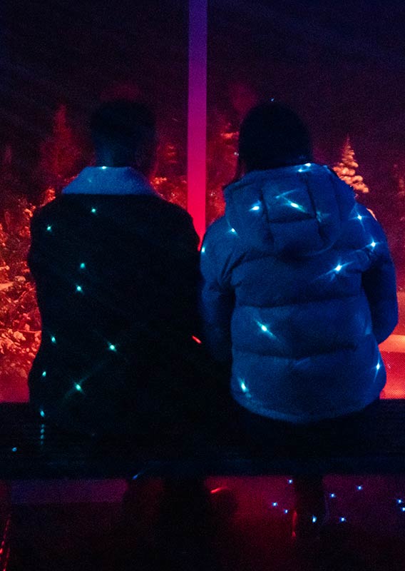 Two people from behind experience Nightrise, with star-shaped lights reflected on their jackets.