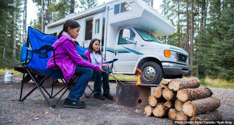 Two kids sitting by a fire pit, beside an RV