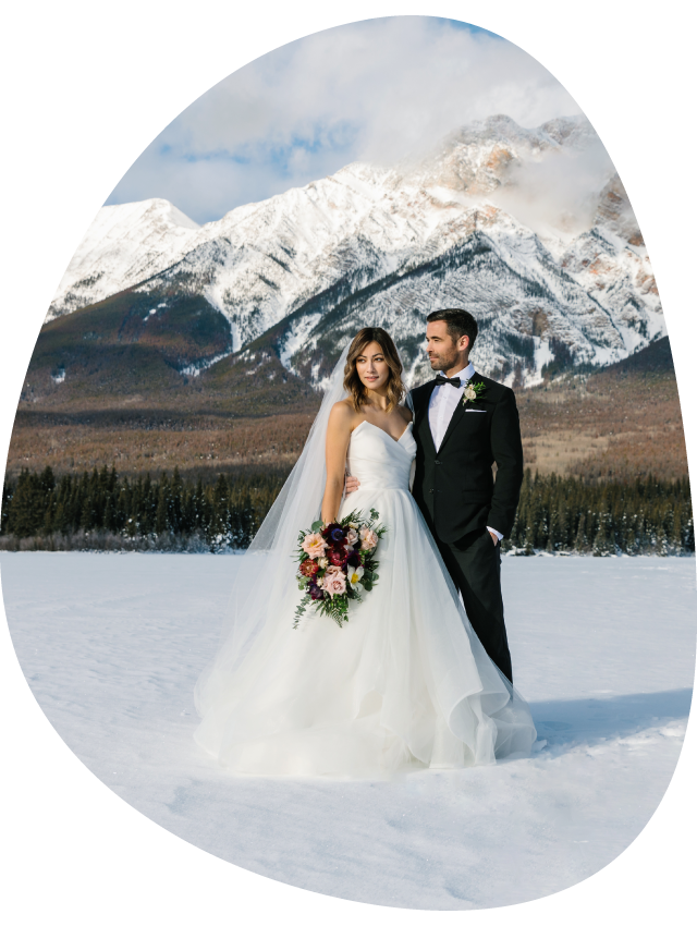 Two brides hold hands at a rocky viewpoint above a small mountain lake.