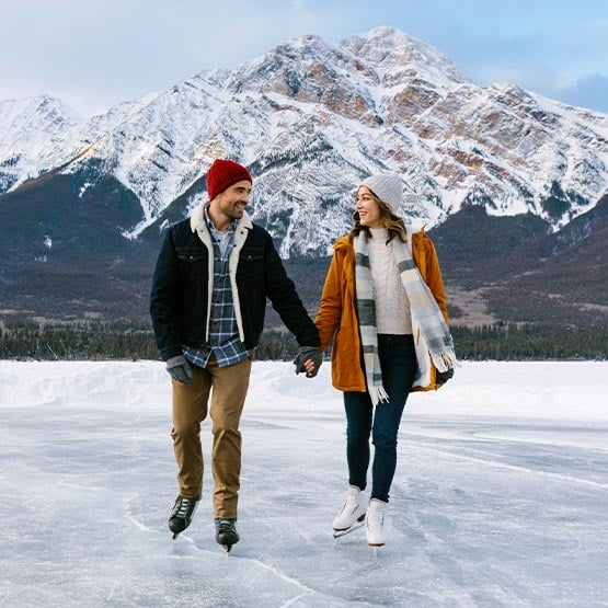 Two people skate on a frozen lake below tall snow-covered mountains.