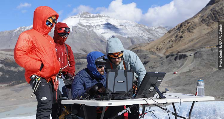 Four researchers look at a laptop on a table set up on the Athabasca Glacier.