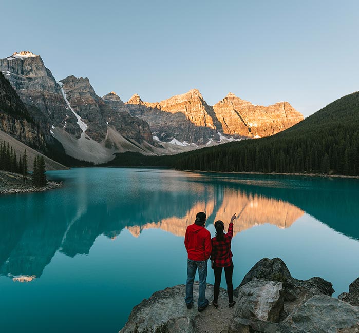 Two people look out from a viewpoint across a blue lake and tall mountains.