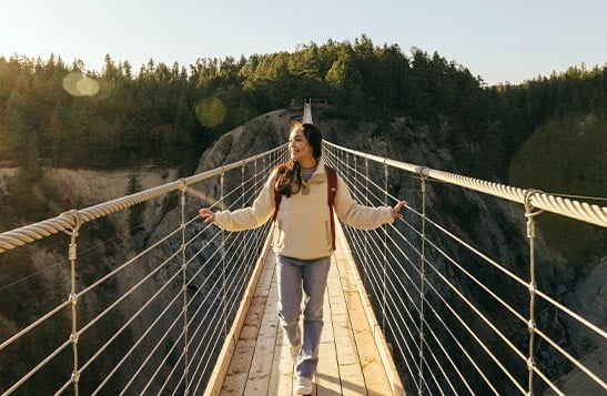 Two people walk across a wooden skybridge overlooking a canyon and trees.