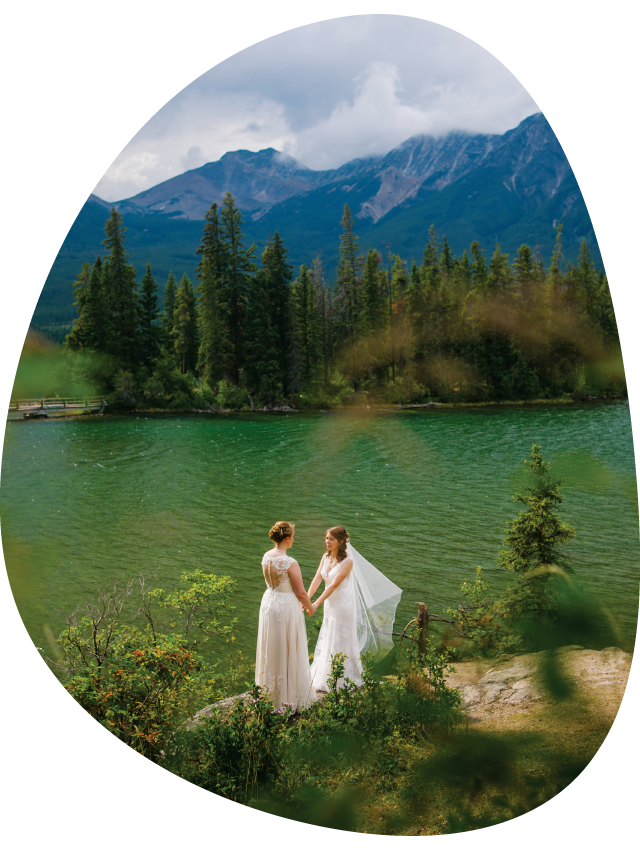 Two brides hold hands at a rocky viewpoint above a small mountain lake.