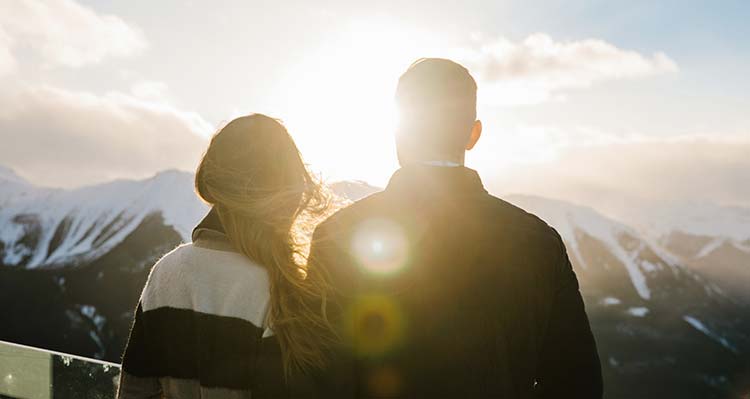 A couple stand at the observation deck with a sunset between them.