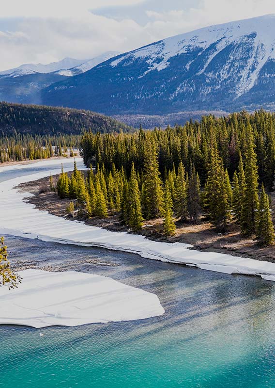 A view of a wide river, flanked by conifer forests and tall, snow-covered mountains.